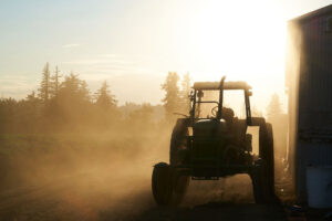 a tractor in near-silhouette against a dramatically lit sky with dust glowing in the setting sunlight. The tractor is next to a structure on the right, and a farm field and distant trees are visible to the left