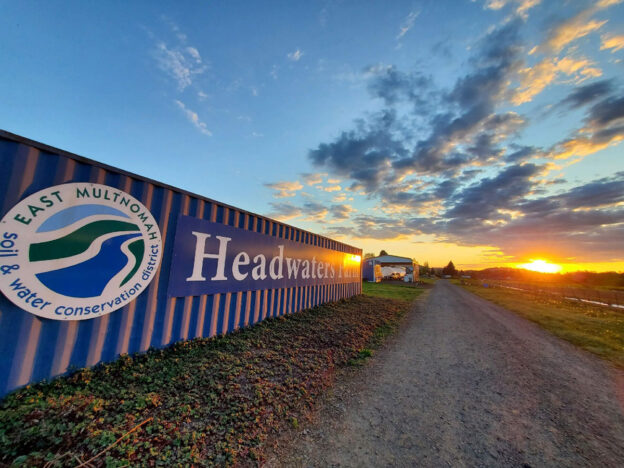 a photo of the sunrise at Headwaters Farm. On the left, a corrugated tool shed wall with a large EMSWCD logo and text "Headwaters Farm," on the right, the farm, the sunrise and a sky with a few scattered clouds lit by the sunrise