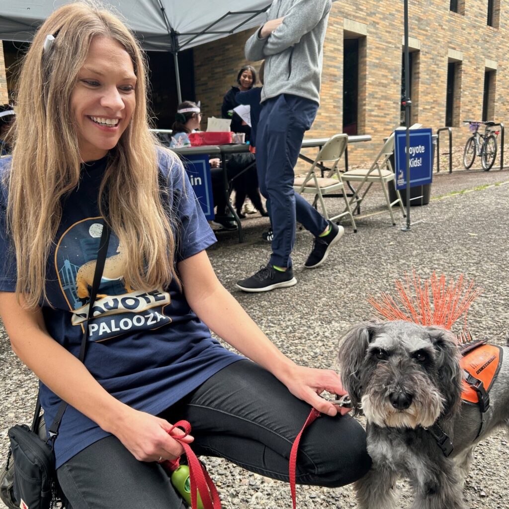 Blonde woman in a blue t-shirt that says Coyote Palooza kneels with a small fluffy dog.