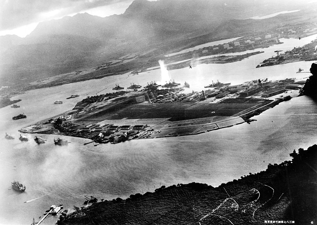 Photograph taken from a Japanese plane during the torpedo attack on ships moored on both sides of Ford Island. View looks about east, with the supply depot, submarine base and fuel tank farm in the right center distance. A torpedo has just hit USS West Virginia on the far side of Ford Island (center). Other battleships moored nearby are (from left): Nevada, Arizona, Tennessee (inboard of West Virginia), Oklahoma (torpedoed and listing) alongside Maryland, and California. On the near side of Ford Island, to the left, are light cruisers Detroit and Raleigh, target and training ship Utah and seaplane tender Tangier. Raleigh and Utah have been torpedoed, and Utah is listing sharply to port. Japanese planes are visible in the right center (over Ford Island) and over the Navy Yard at right. Japanese writing in the lower right states that the photograph was reproduced by authorization of the Navy Ministry. U.S. Naval History and Heritage Command Photograph.